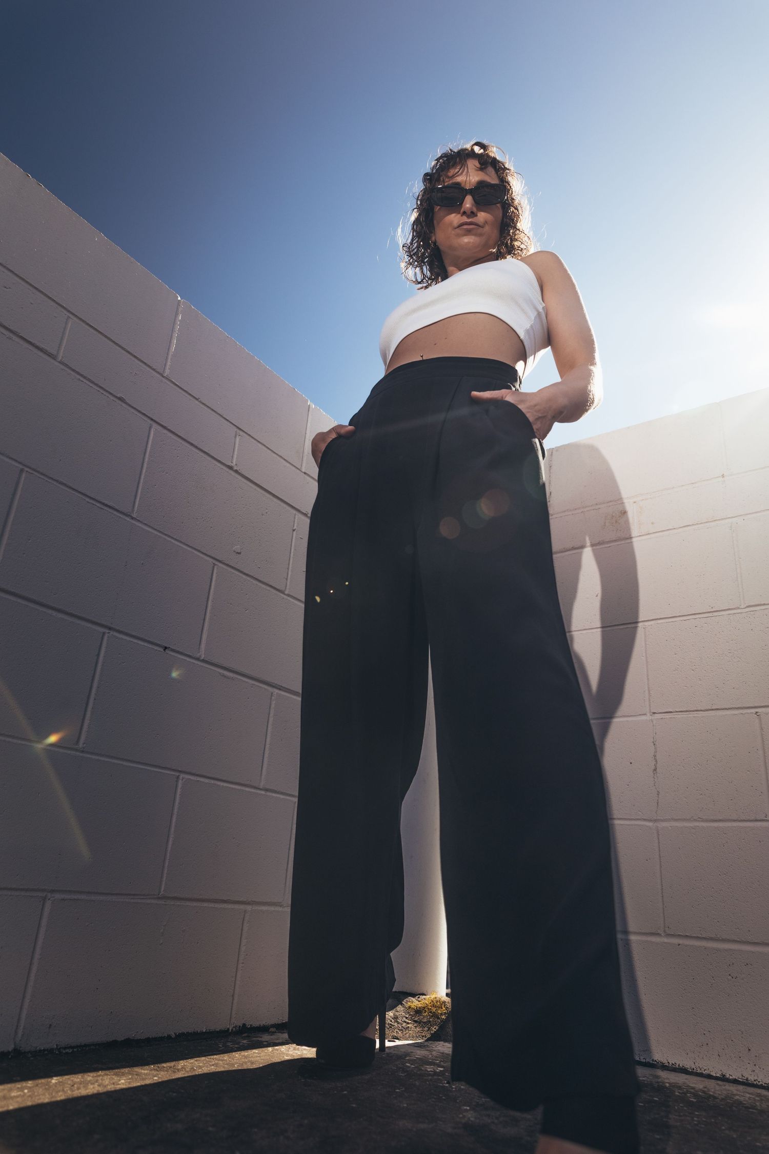Stylish woman in white crop top and black wide-leg pants stands against white brick wall in bright sunlight.