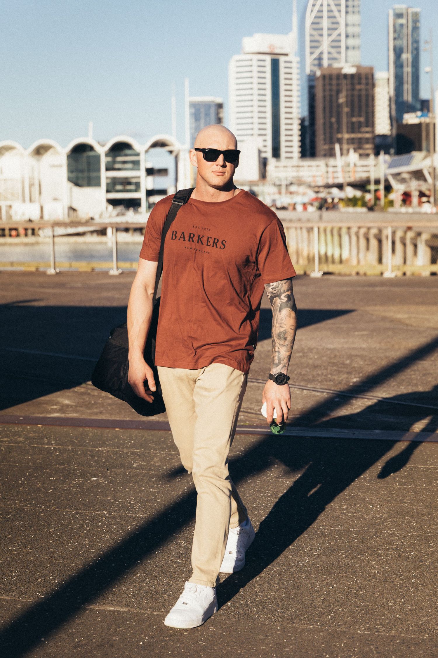Bald tattooed man in brown Barkers t-shirt, khaki pants, white sneakers, carrying bag, walking in urban cityscape.