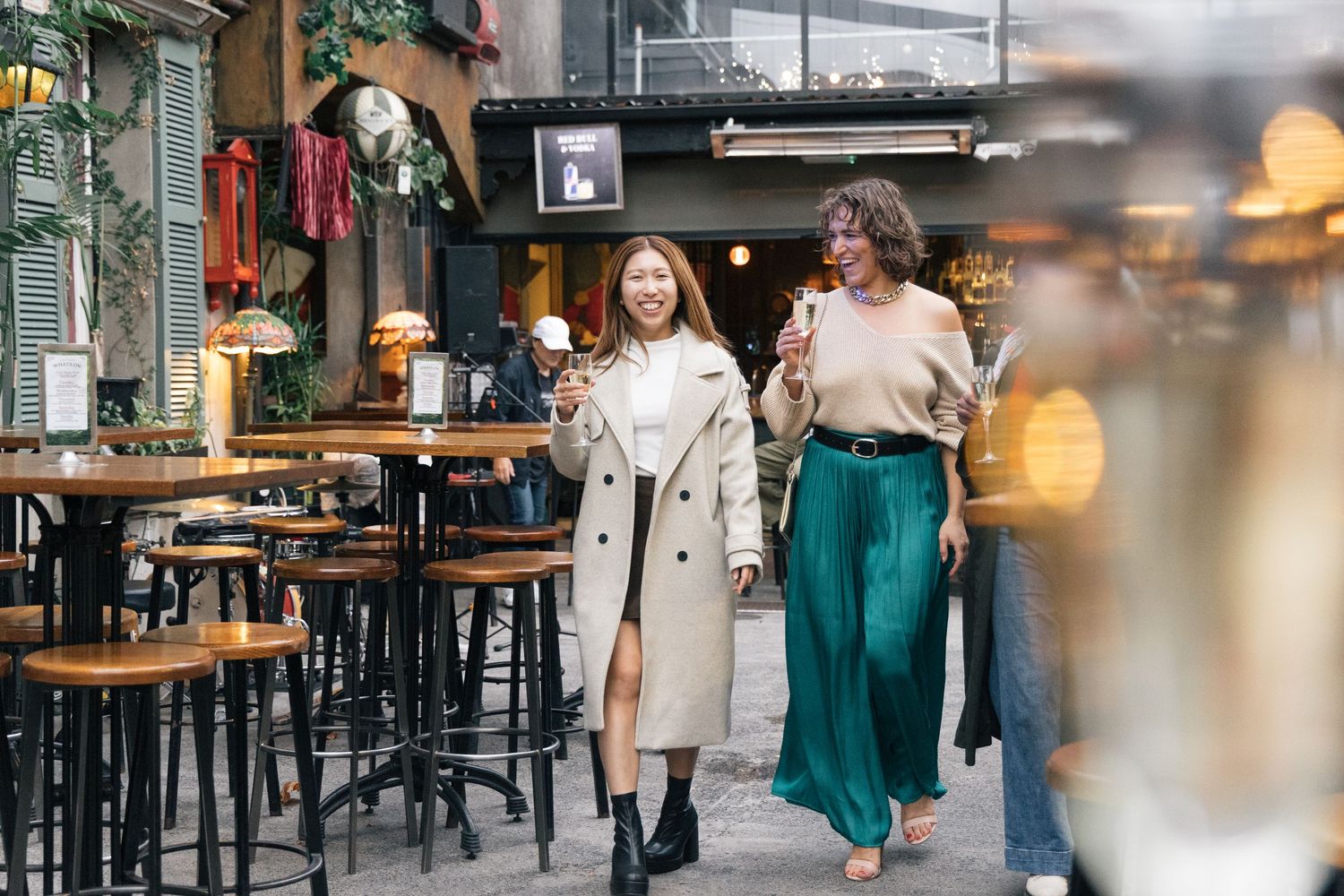Two stylish women walking and smiling outside a trendy cafe bar holding drinks.
