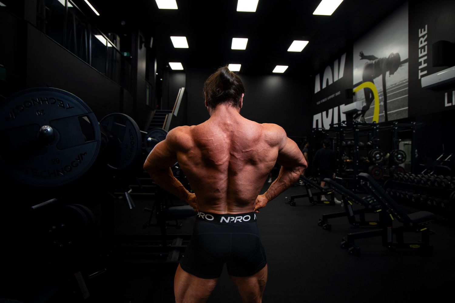 Muscular bodybuilder posing back muscles in dark gym wearing NARO shorts near barbell rack.