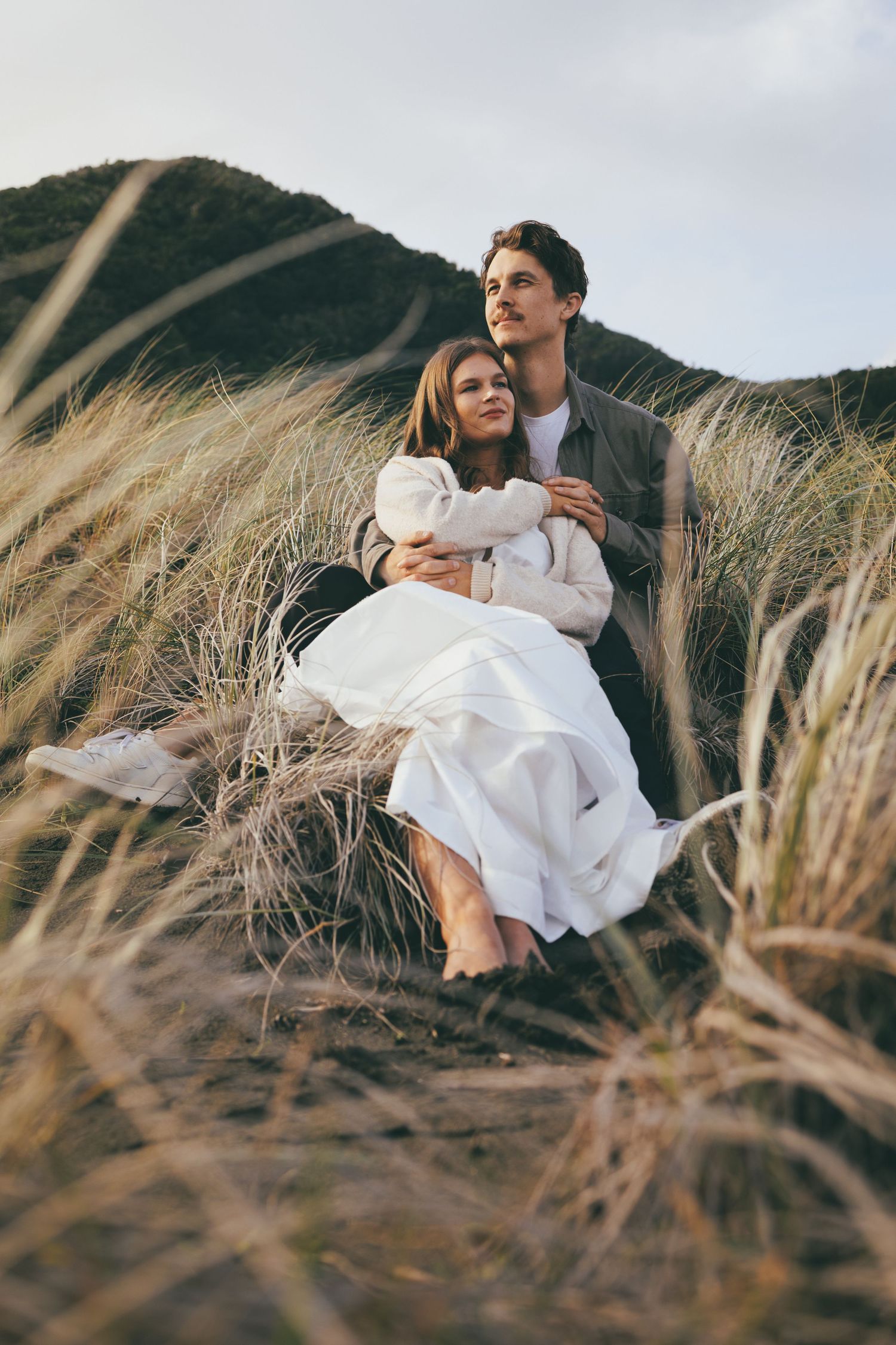 Couple embracing in tall windswept beach grass with dramatic hills in the background.