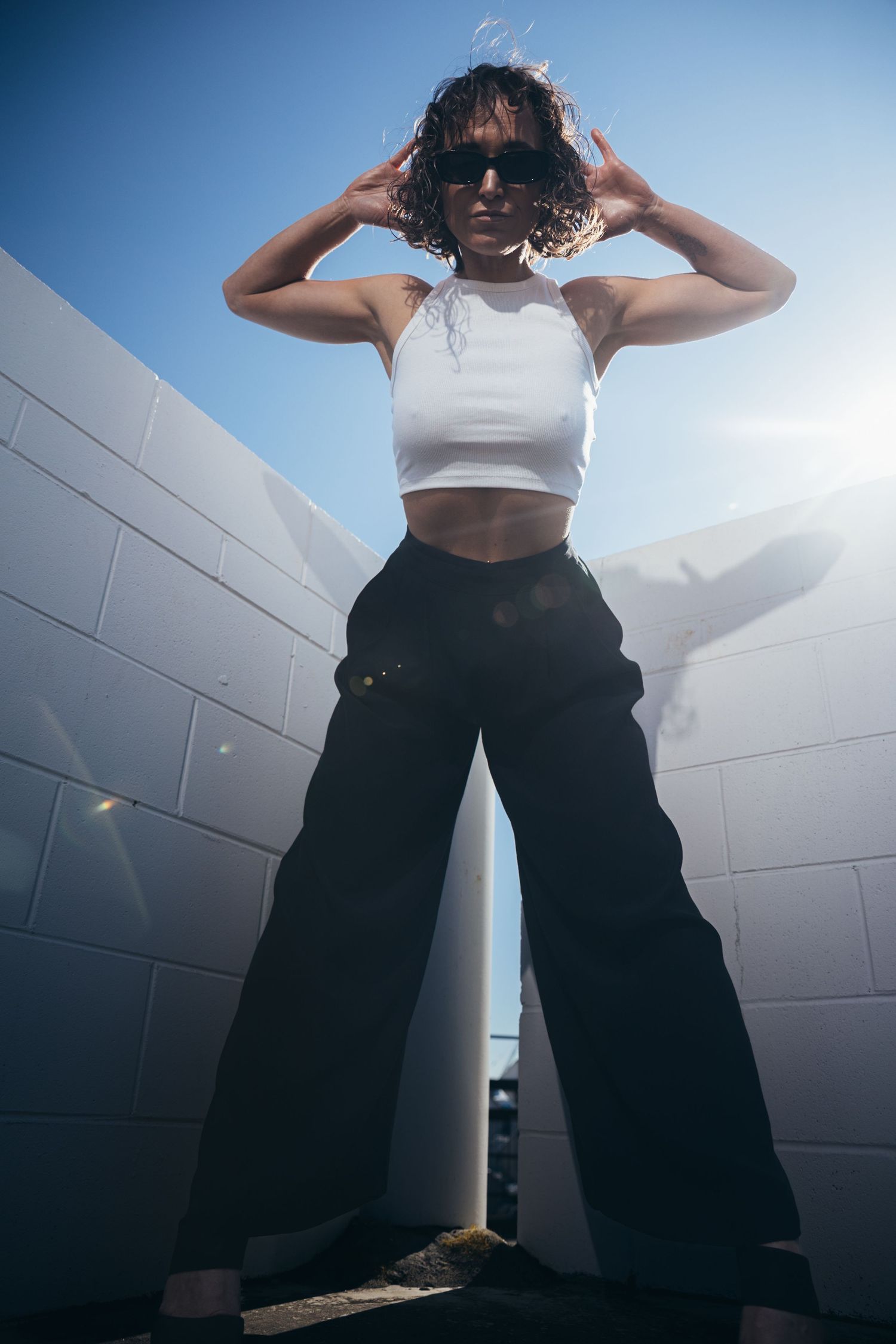 Confident woman in white crop top and black wide-leg pants posing with hands on head against sunny blue sky.