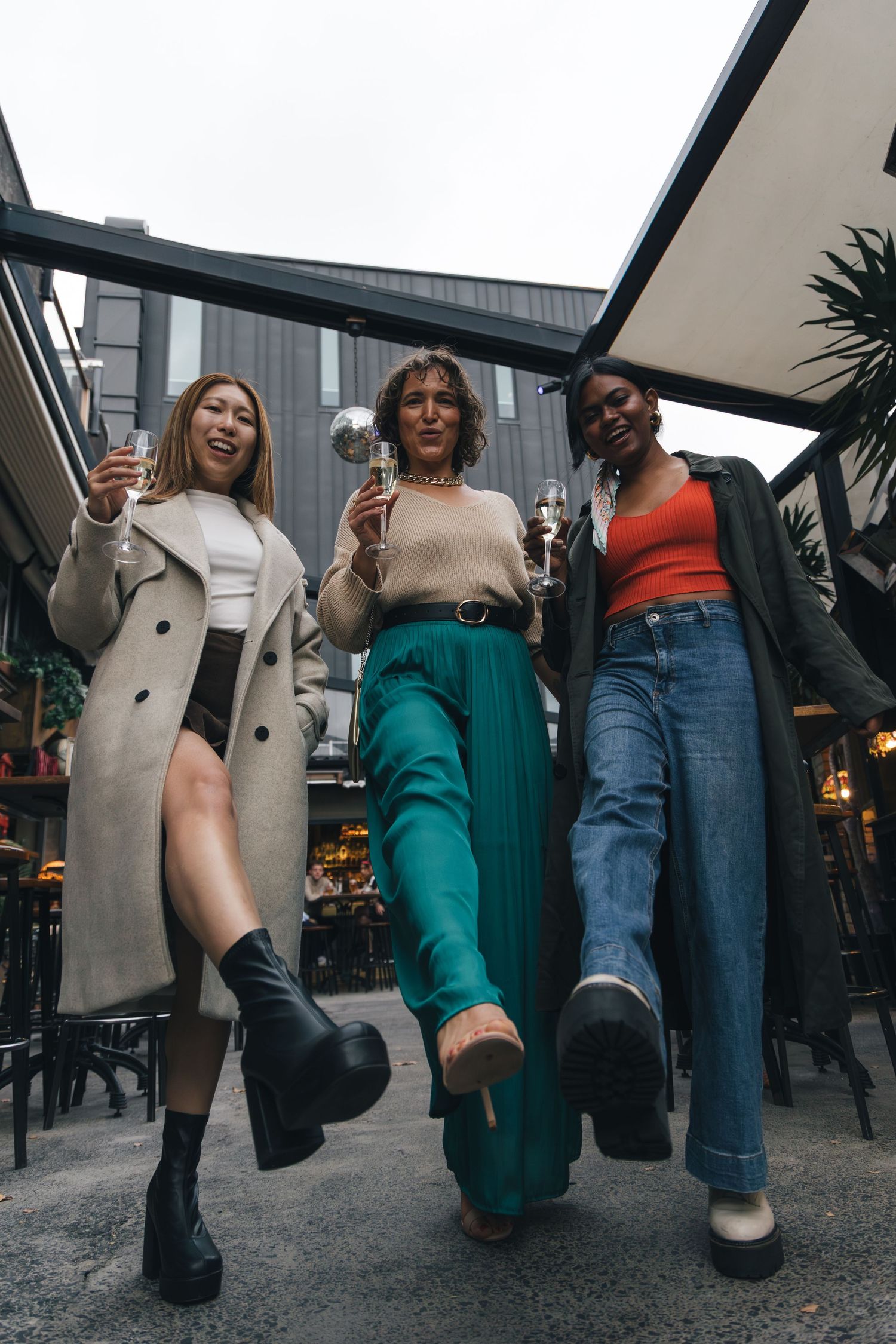Three stylish women walk confidently holding cocktails at a trendy outdoor bar venue.