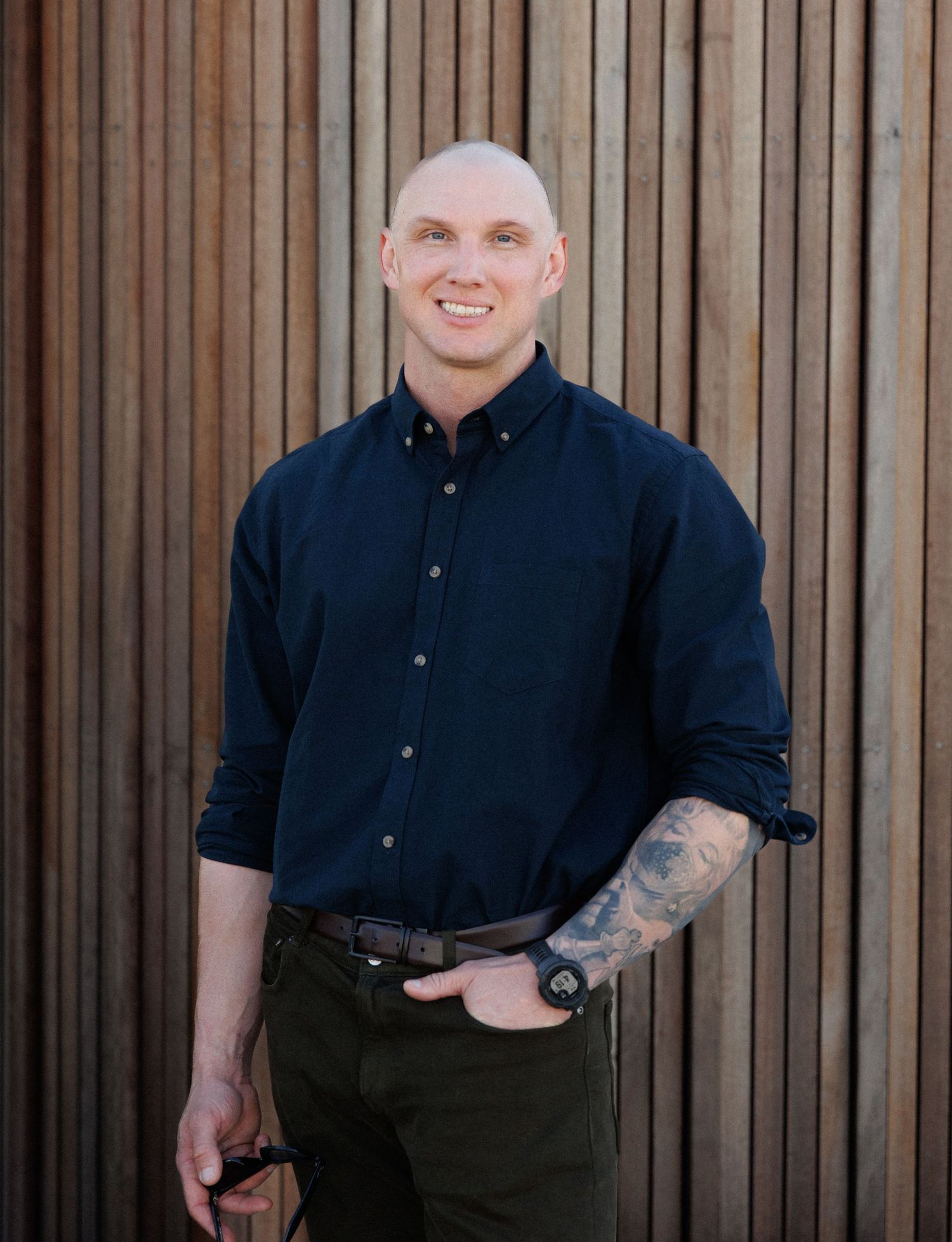 Smiling bald man with tattoos wearing a navy shirt, posing casually against a wooden slat wall backdrop.