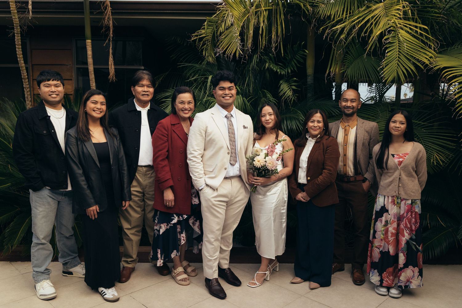 Wedding couple poses with family group photo in front of tropical palm trees outdoors.