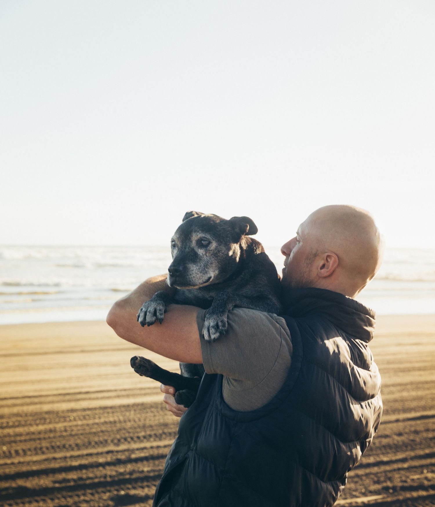Bald man carrying his black dog in arms on sandy beach with ocean waves in background.