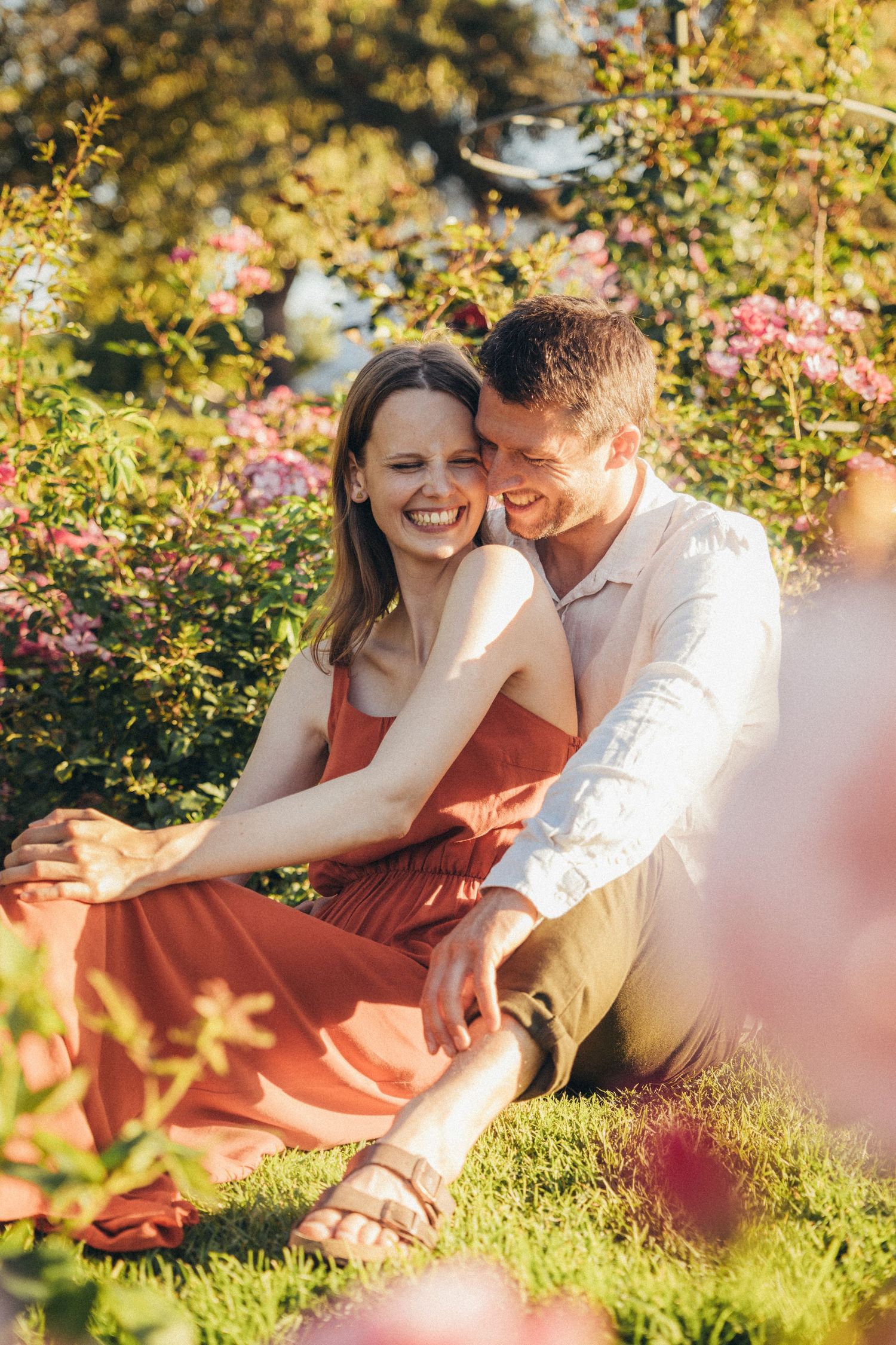 Happy couple sitting on grass surrounded by pink roses, woman in red dress laughing joyfully while man embraces her.