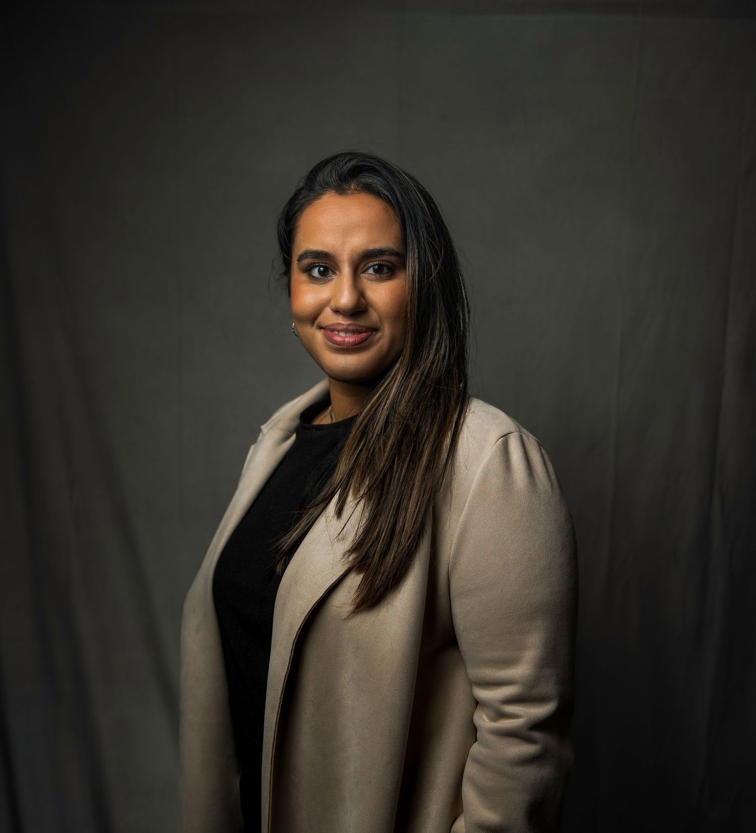 Woman in beige blazer and black top poses smiling against dark fabric backdrop.