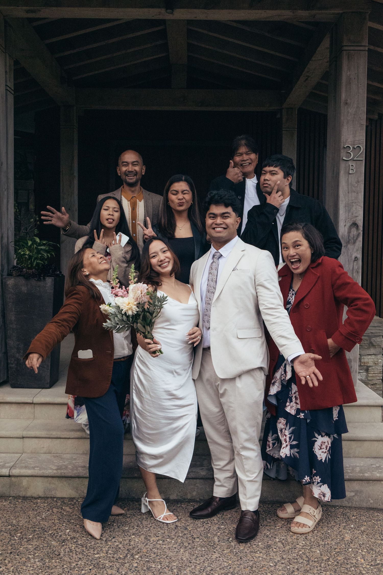 A joyful wedding group photo with a bride in white dress holding bouquet and groom in white suit, surrounded by celebrating family.