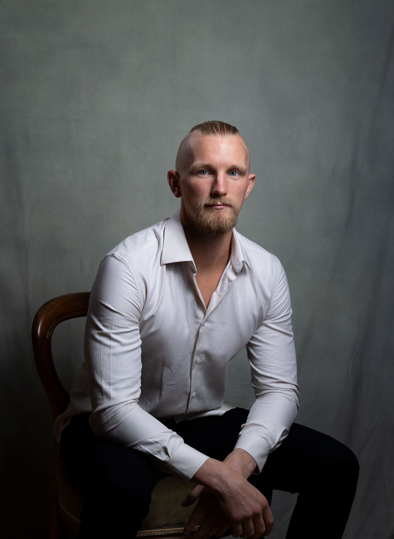 Athletic man in white shirt sitting on wooden chair in moody studio portrait.