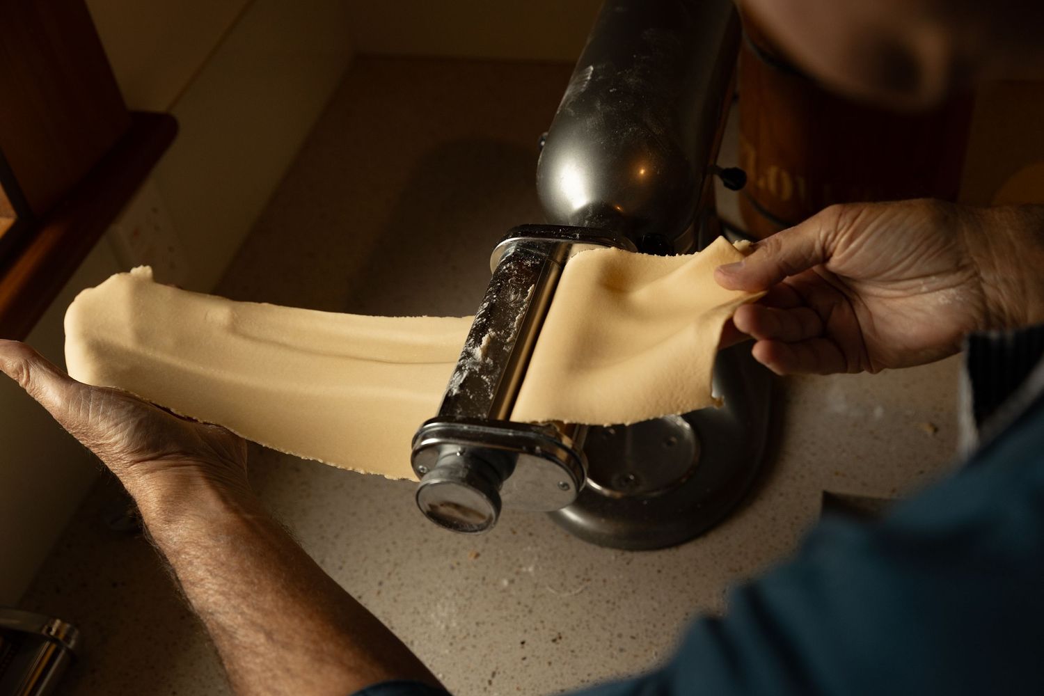 Hands feeding fresh pasta dough through a KitchenAid pasta roller attachment on a countertop.