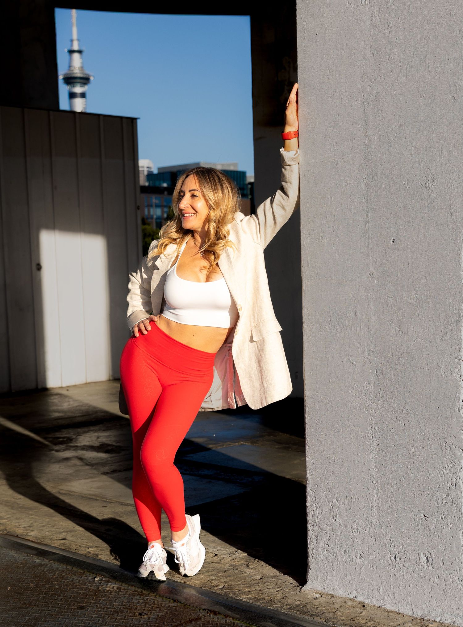 Blonde woman in red leggings and white blazer posing in urban doorway with sky tower.