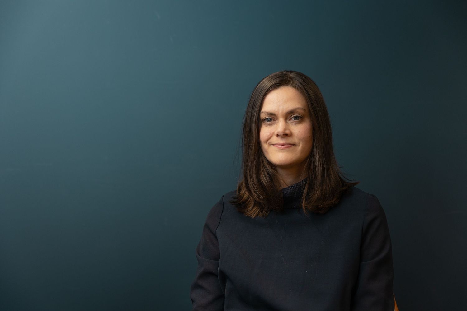 Smiling woman with dark shoulder-length hair wearing a black turtleneck against a teal wall.