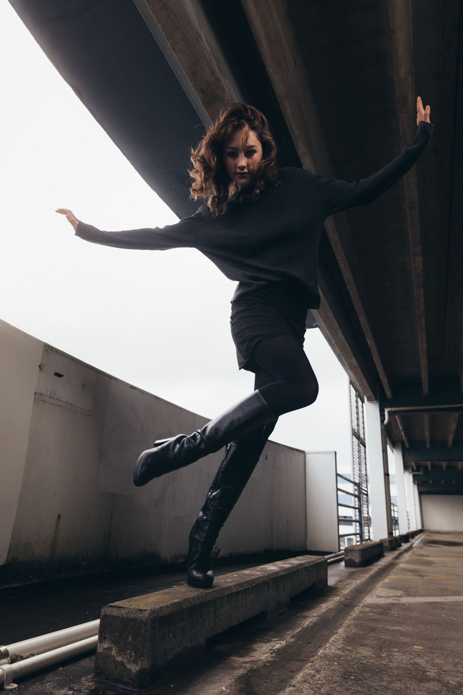 Woman in black outfit balancing on concrete ledge under urban bridge in dramatic dance pose.