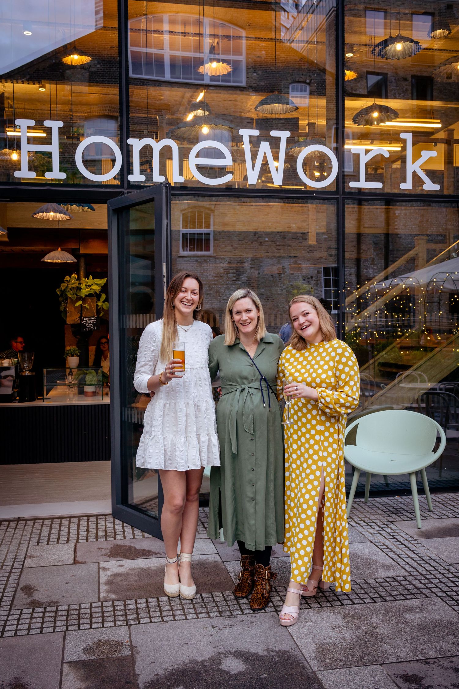 Three women pose smiling outside HomeWork venue, wearing white, green, and yellow polka dot dresses.