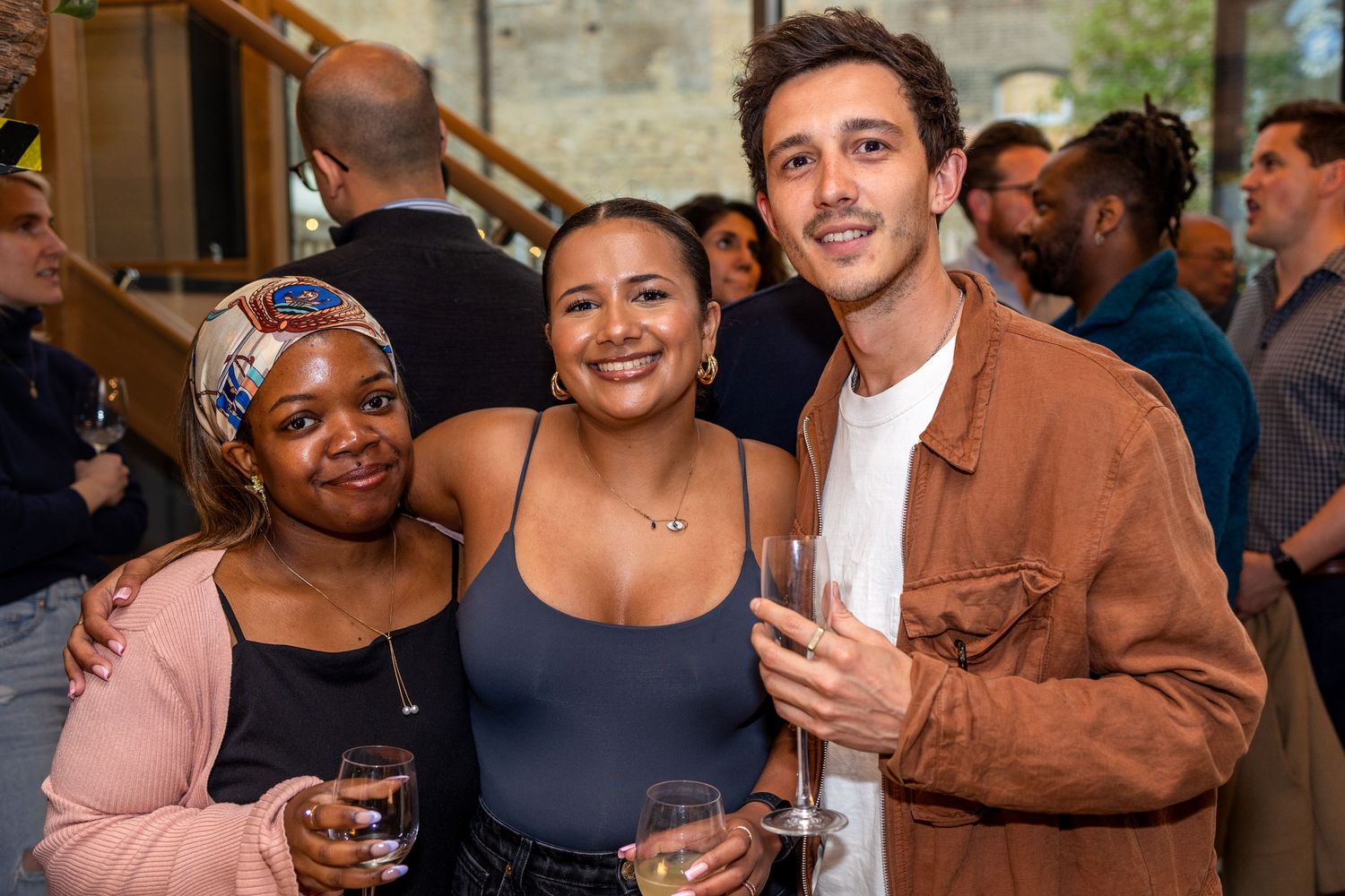 Two women and a man pose together smiling while holding drinks at an indoor gathering.