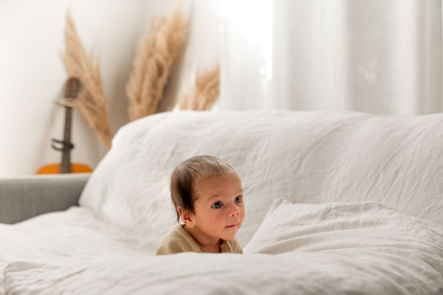 Adorable baby sitting on a white linen bed with soft natural light and dried pampas grass decor in background.