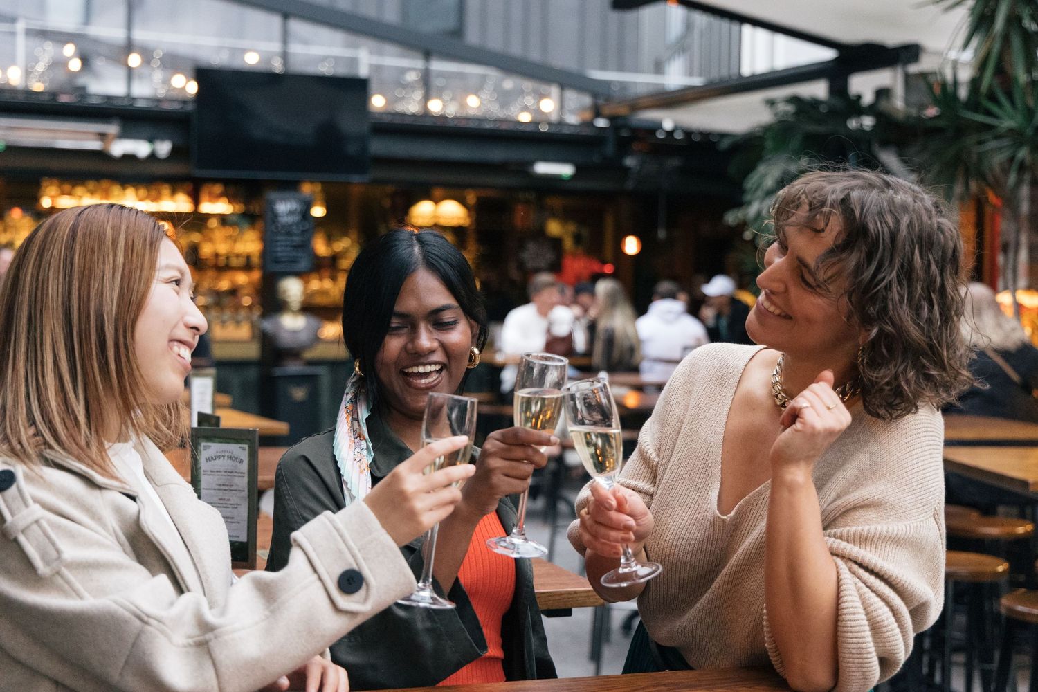 Three diverse women toasting with champagne glasses at a restaurant, smiling and celebrating together.