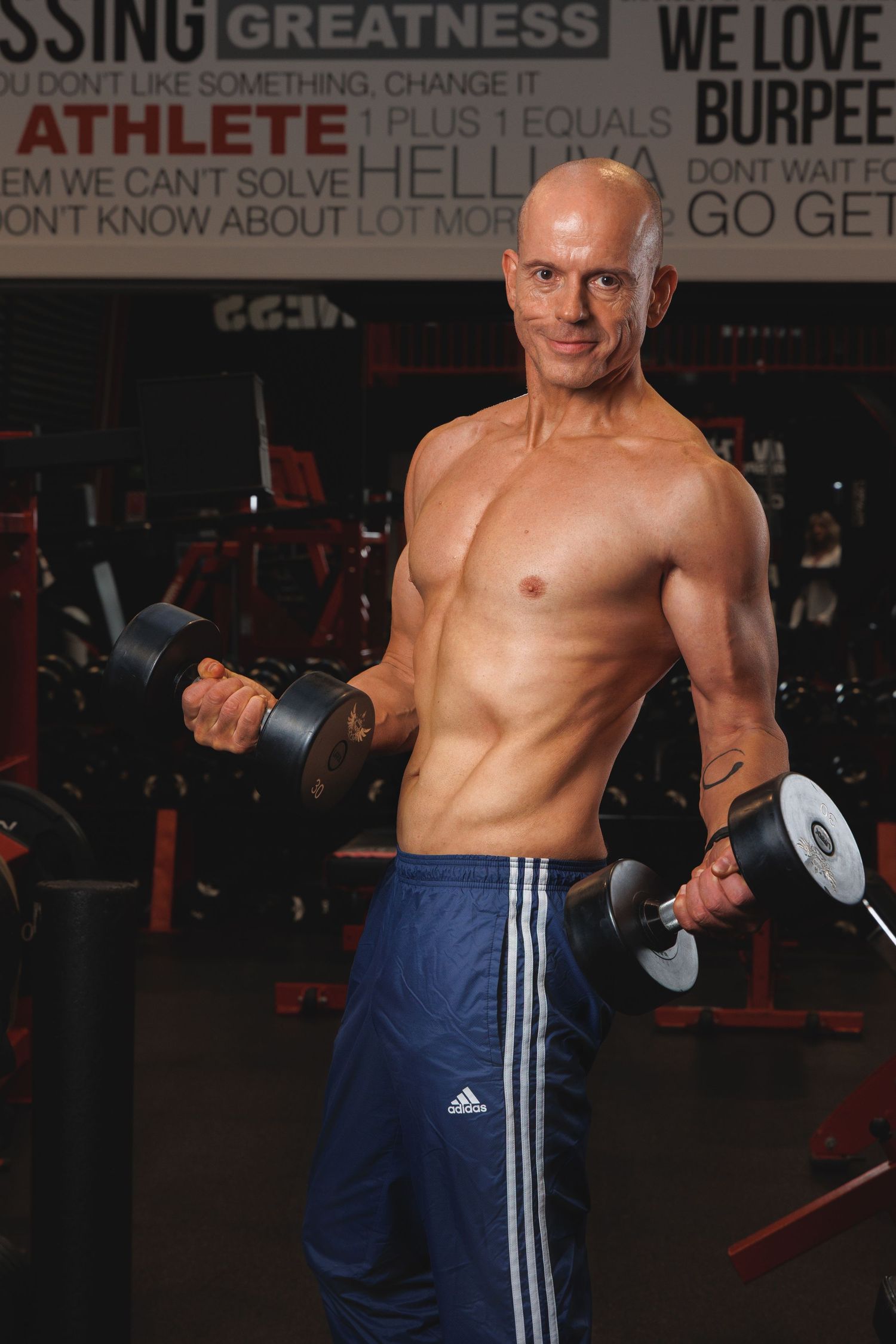 Muscular shirtless man lifting dumbbells in a gym with motivational text on the wall.