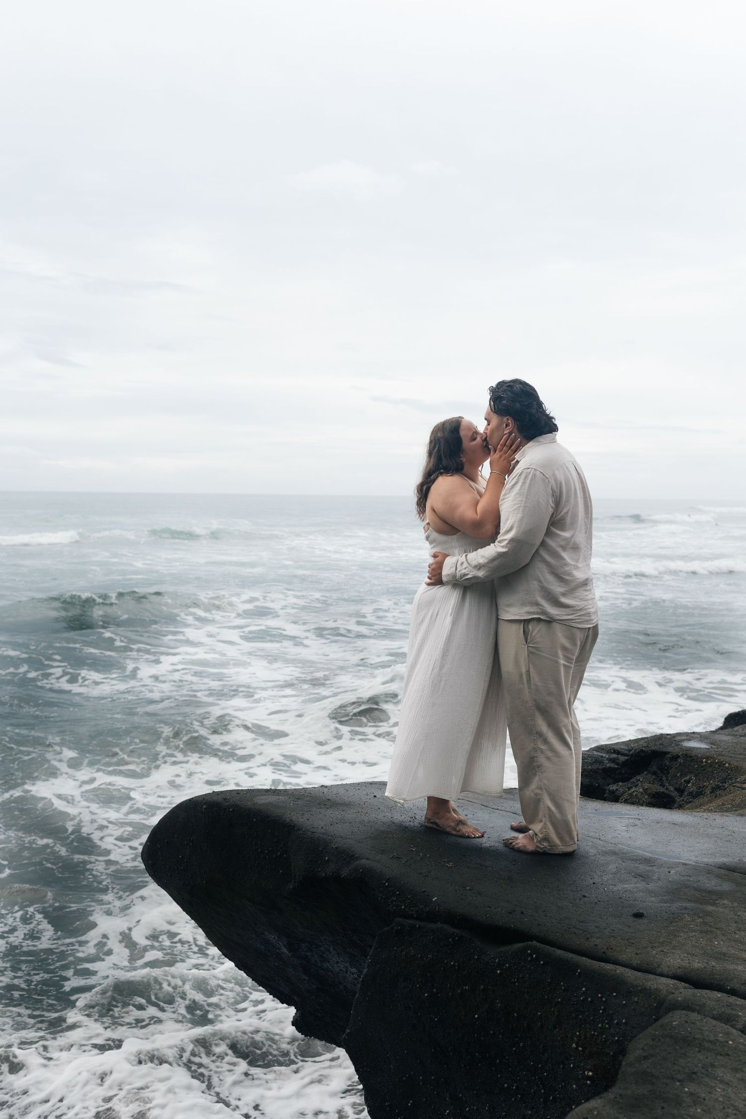 Couple sharing a kiss on a rocky ocean cliff with waves crashing below.