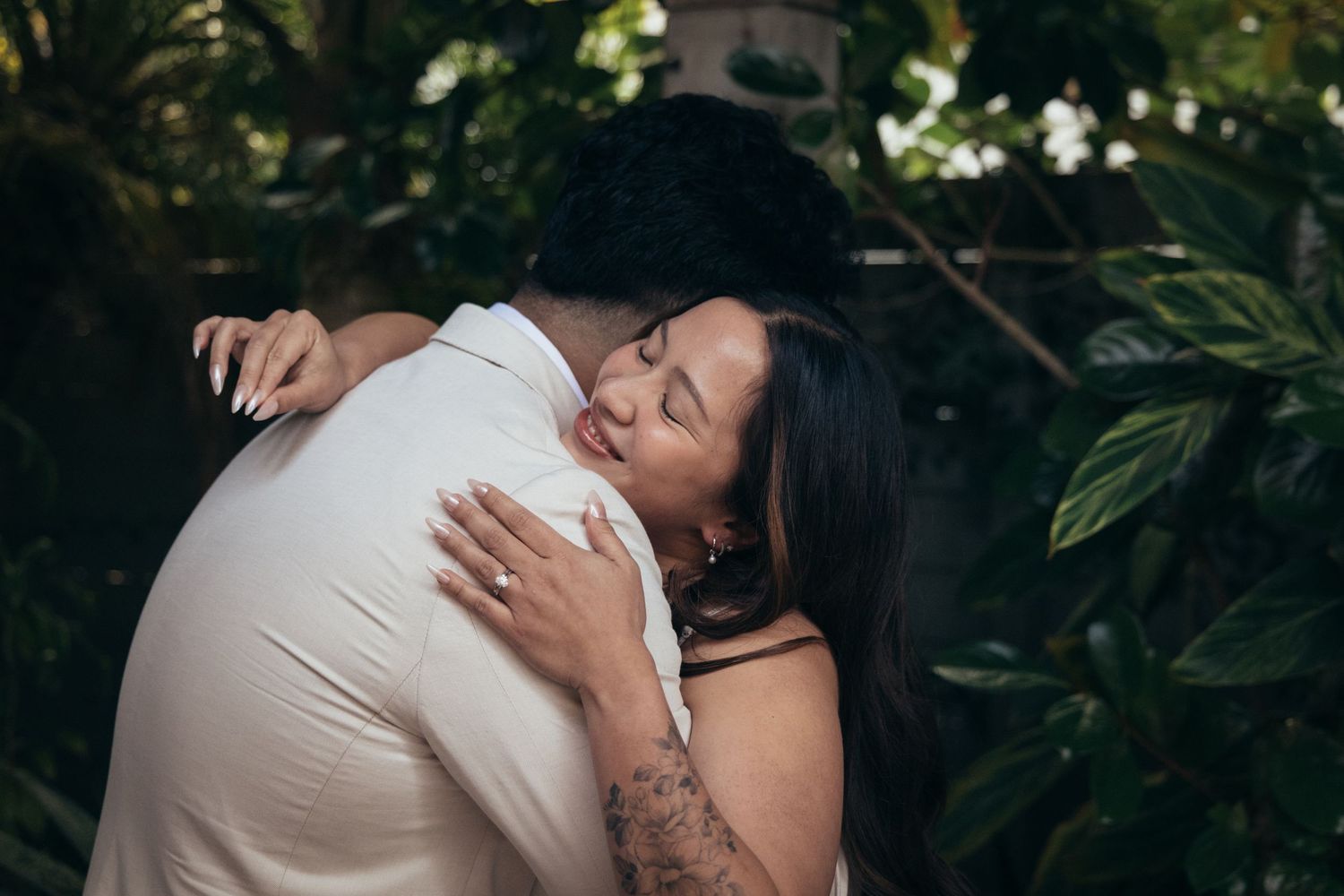 Couple embracing tenderly outdoors among lush green tropical plants, woman with tattoo hugging man in white shirt.