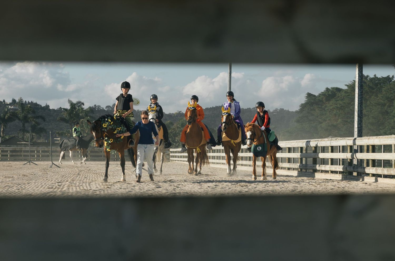 Group of young equestrian riders on horses in sandy arena viewed through wooden fence.