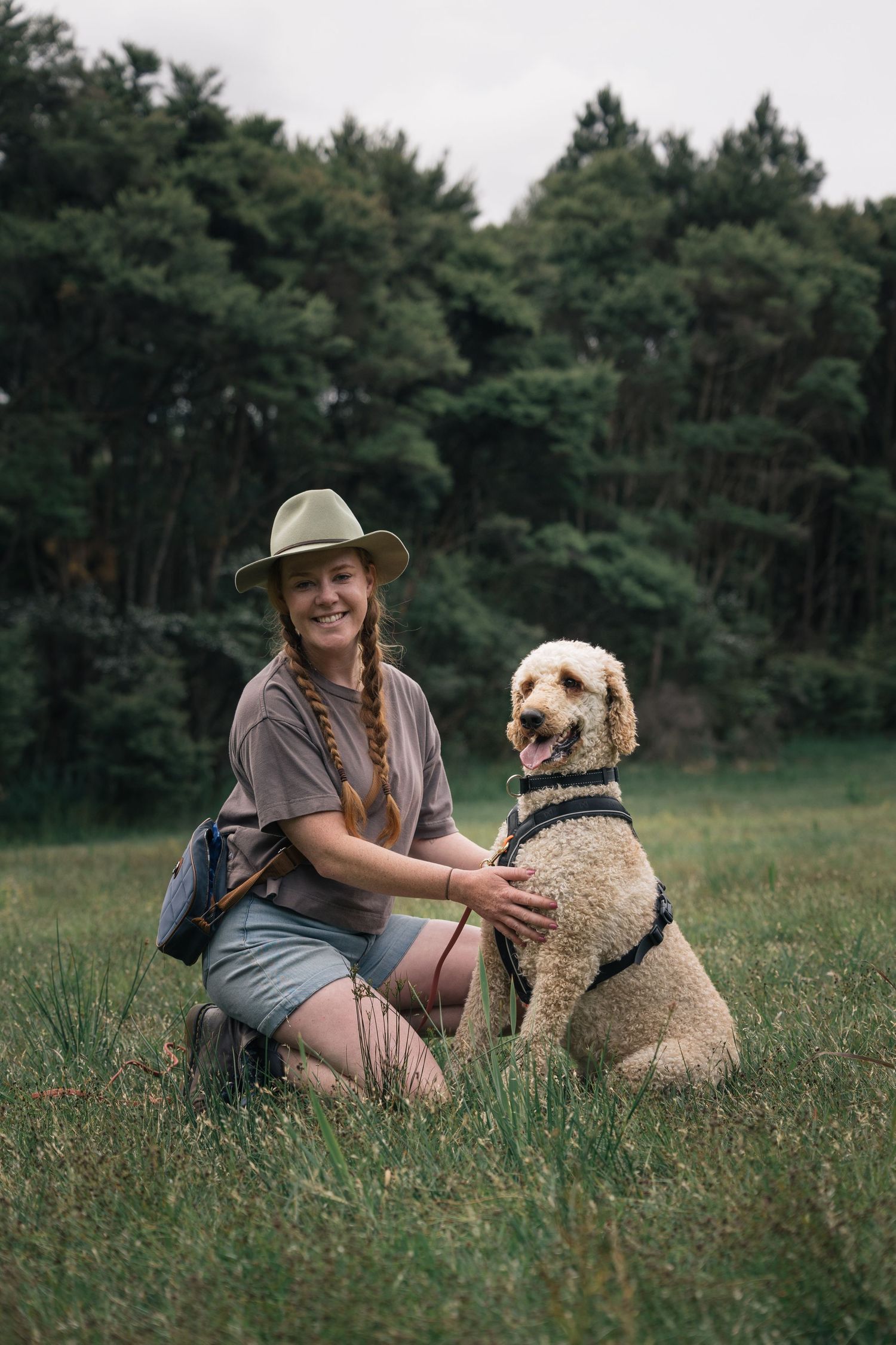 Happy woman in hat kneeling beside golden labradoodle dog sitting in green grassy field outdoors.