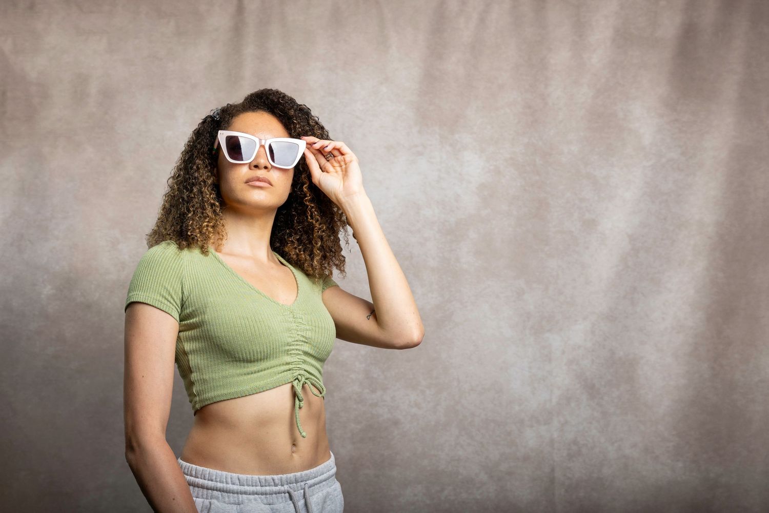 Woman with curly hair in green crop top adjusting white sunglasses against grey backdrop.