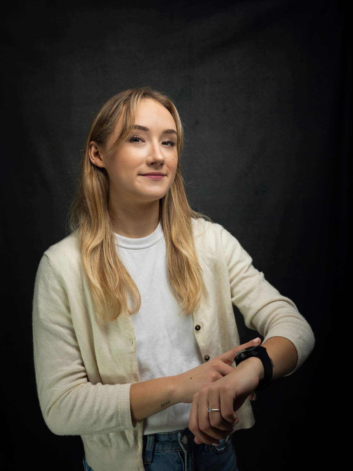 Young blonde woman in cream cardigan checking smartwatch against dark studio background.