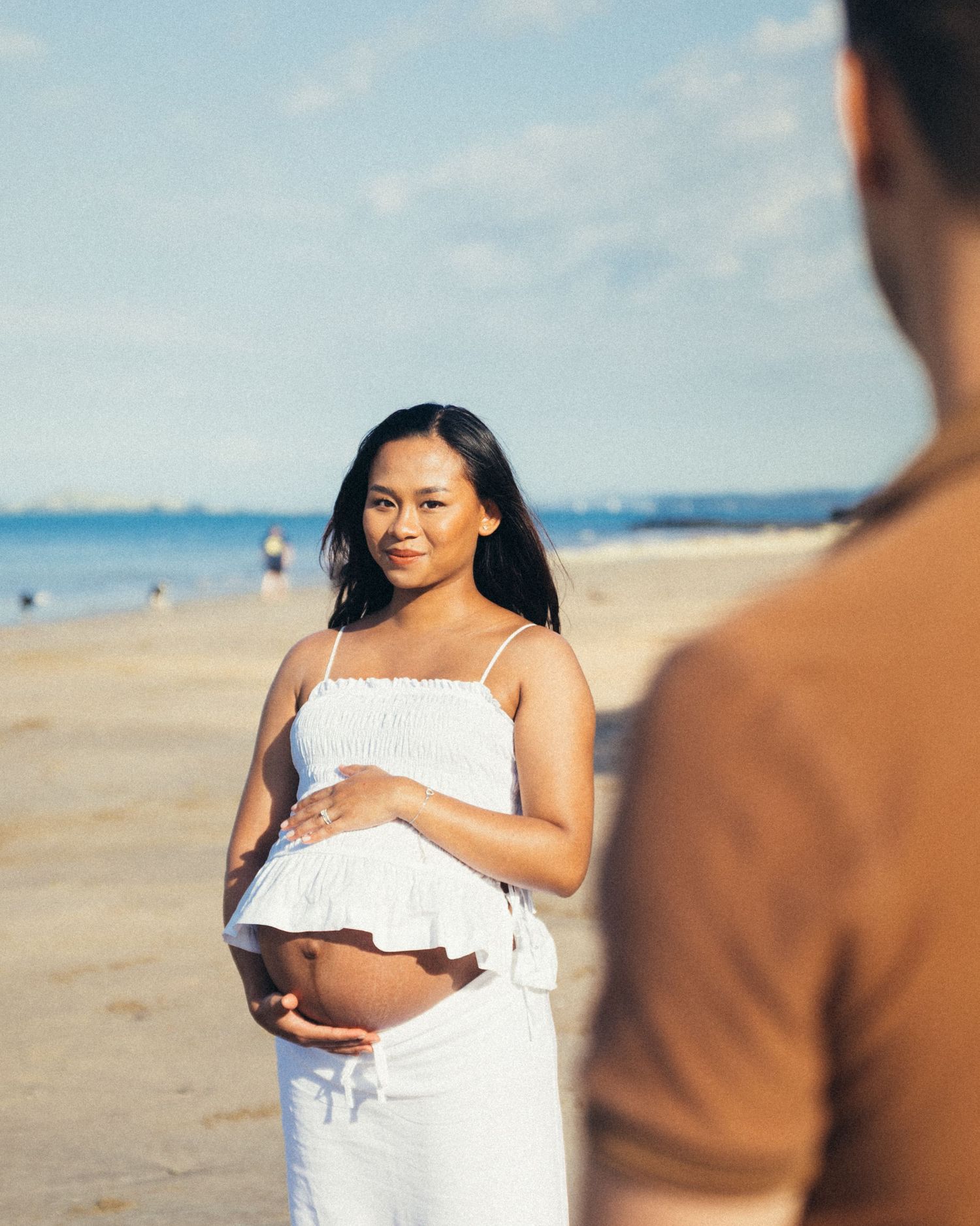 Pregnant woman in white outfit smiling on a sunny beach, cradling her baby bump during a maternity photoshoot.