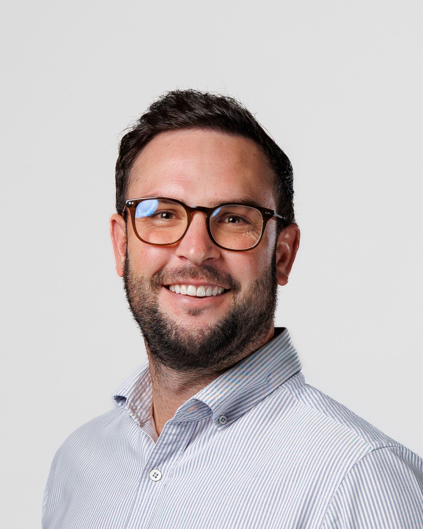 Professional headshot of a smiling man with dark hair and beard wearing glasses and a striped shirt.