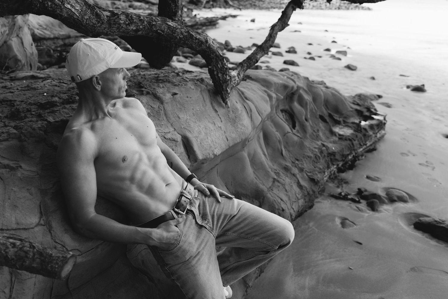 Fit shirtless man resting on rocks at a beach in black and white photo wearing a cap.