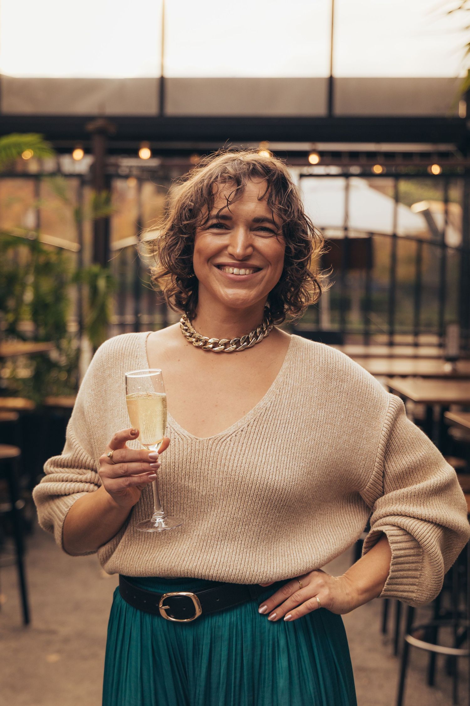 Smiling woman holding champagne glass wearing beige sweater and green skirt with gold chain necklace at a restaurant.