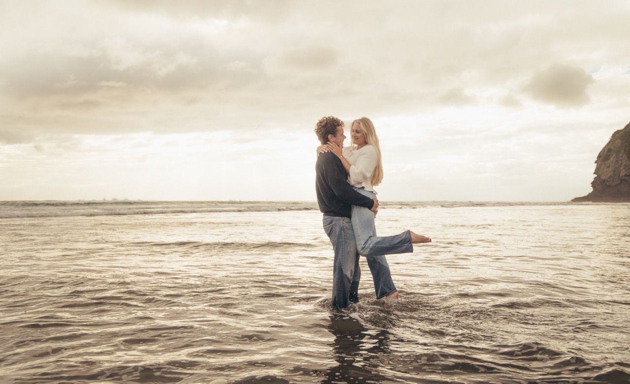 Romantic couple embracing in ocean waves at sunset on a scenic beach with dramatic cloudy skies.