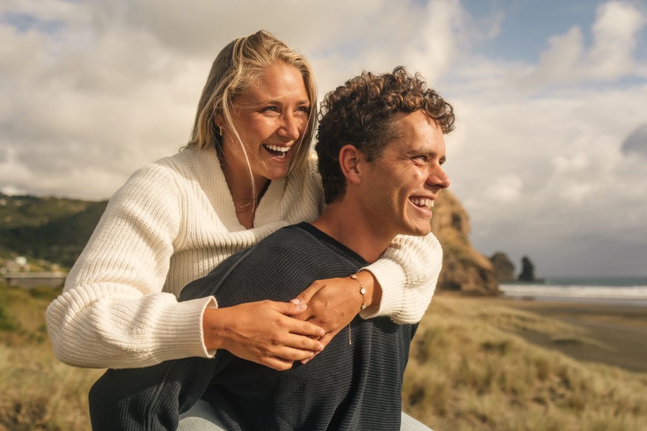 A happy couple laughs together on a beach, woman in white sweater giving a piggyback ride near rocky coastline.