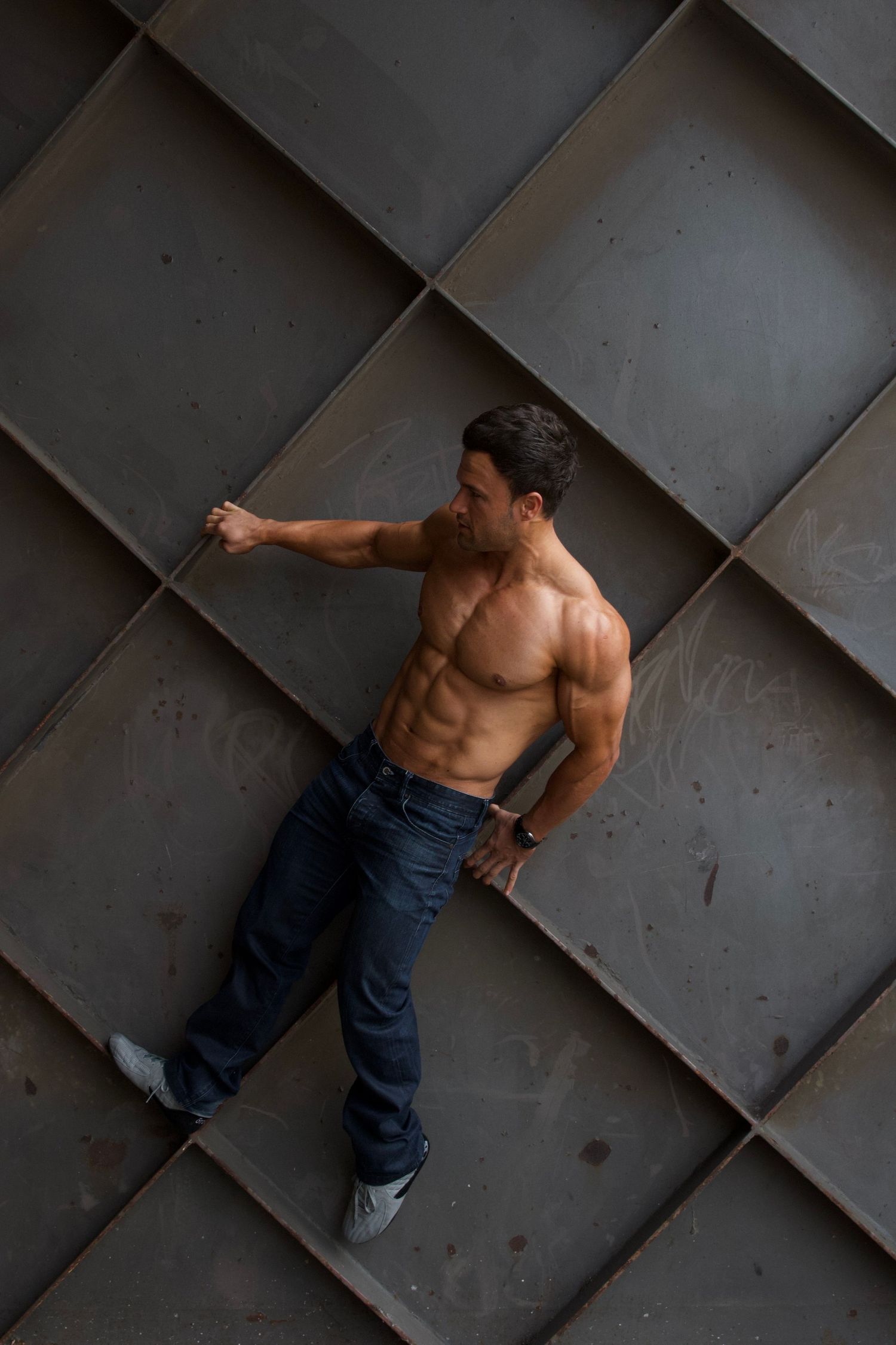 Shirtless muscular man in jeans leaning against a geometric metal wall panel background.