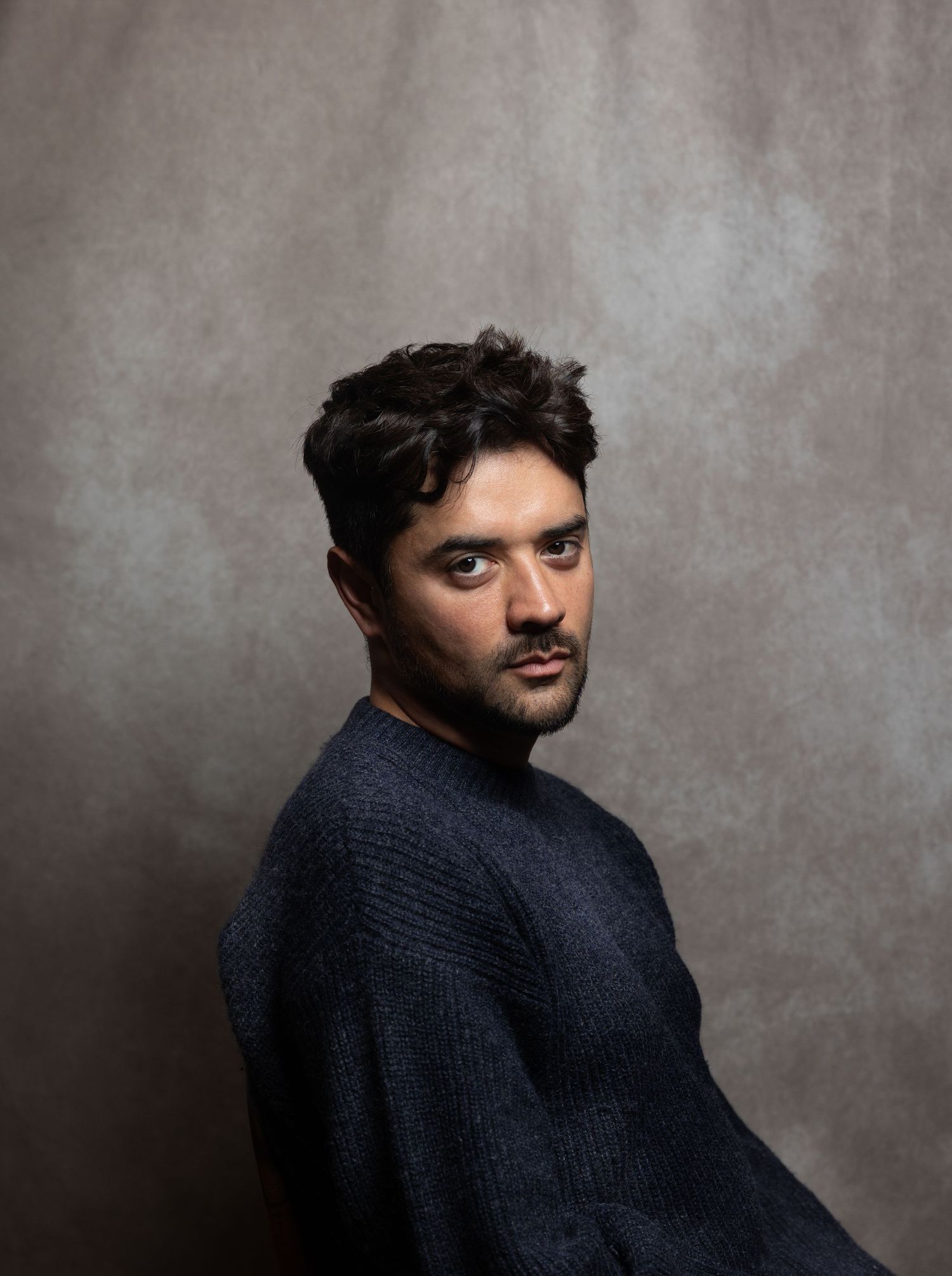 Dark-haired man in navy sweater giving an intense look over shoulder against gray background.