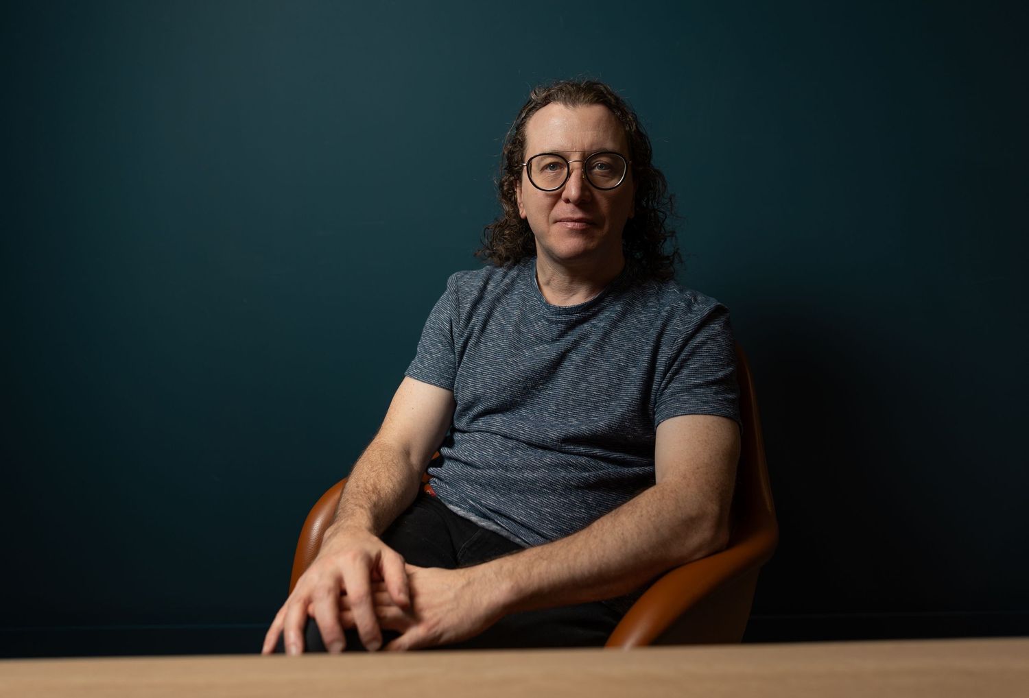 Man with curly hair and glasses in a gray t-shirt seated at a wooden table against a teal wall.
