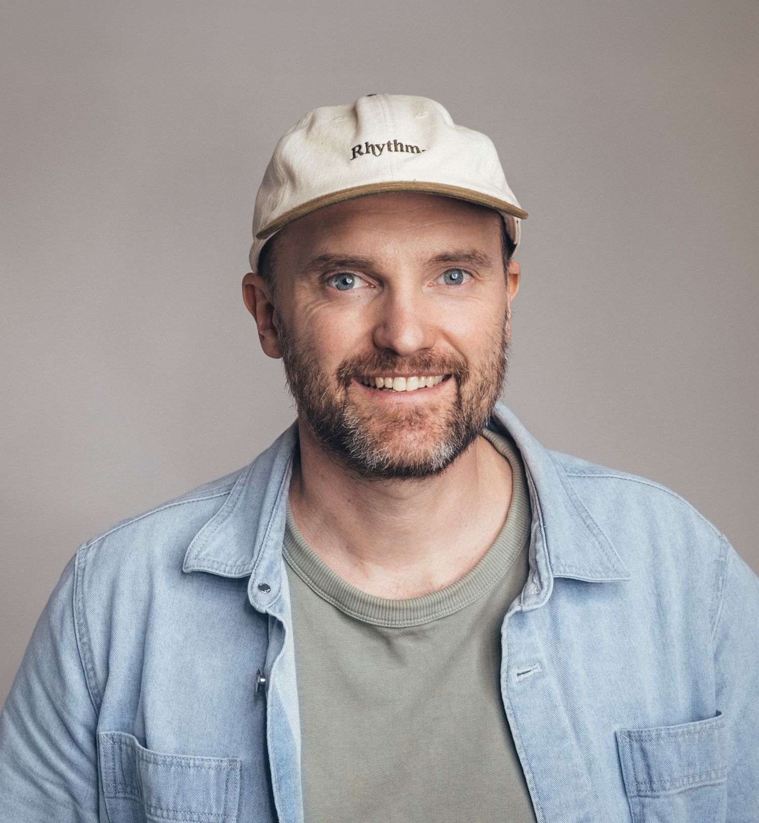 Smiling bearded man wearing a cream Rhythm cap and blue shirt against a grey background.