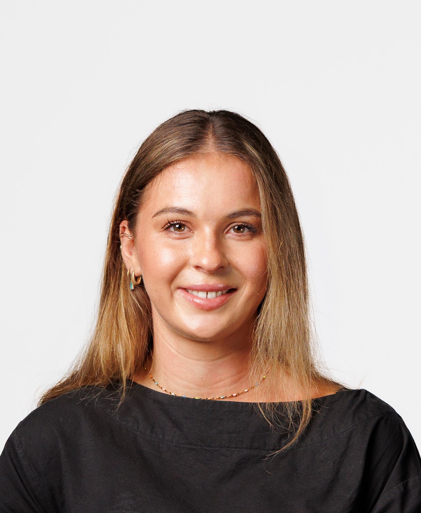 Young woman with blonde hair smiling, wearing a black top with gold necklace against a light grey background.