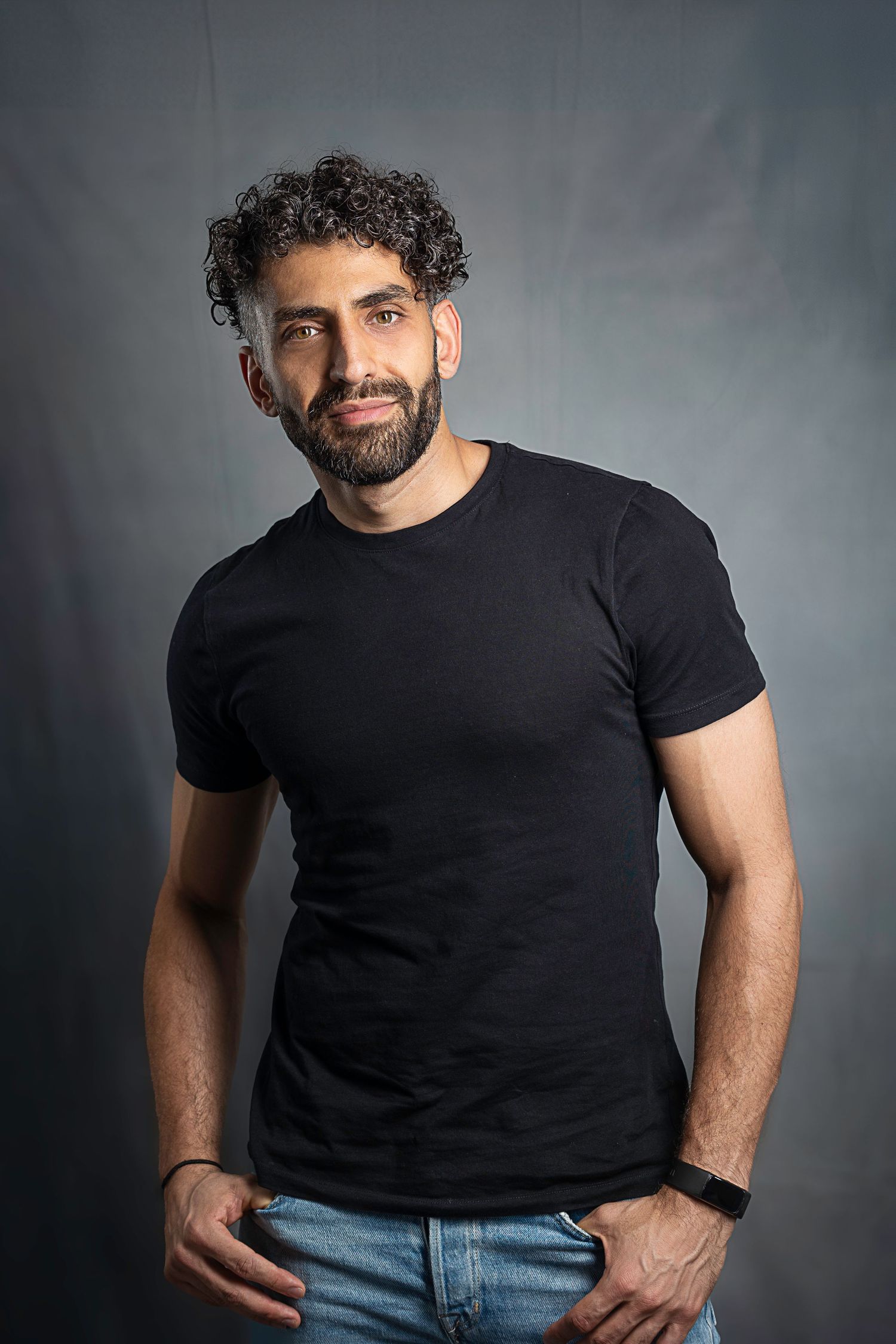 Fit man with curly hair and beard in black t-shirt and jeans posing in studio.