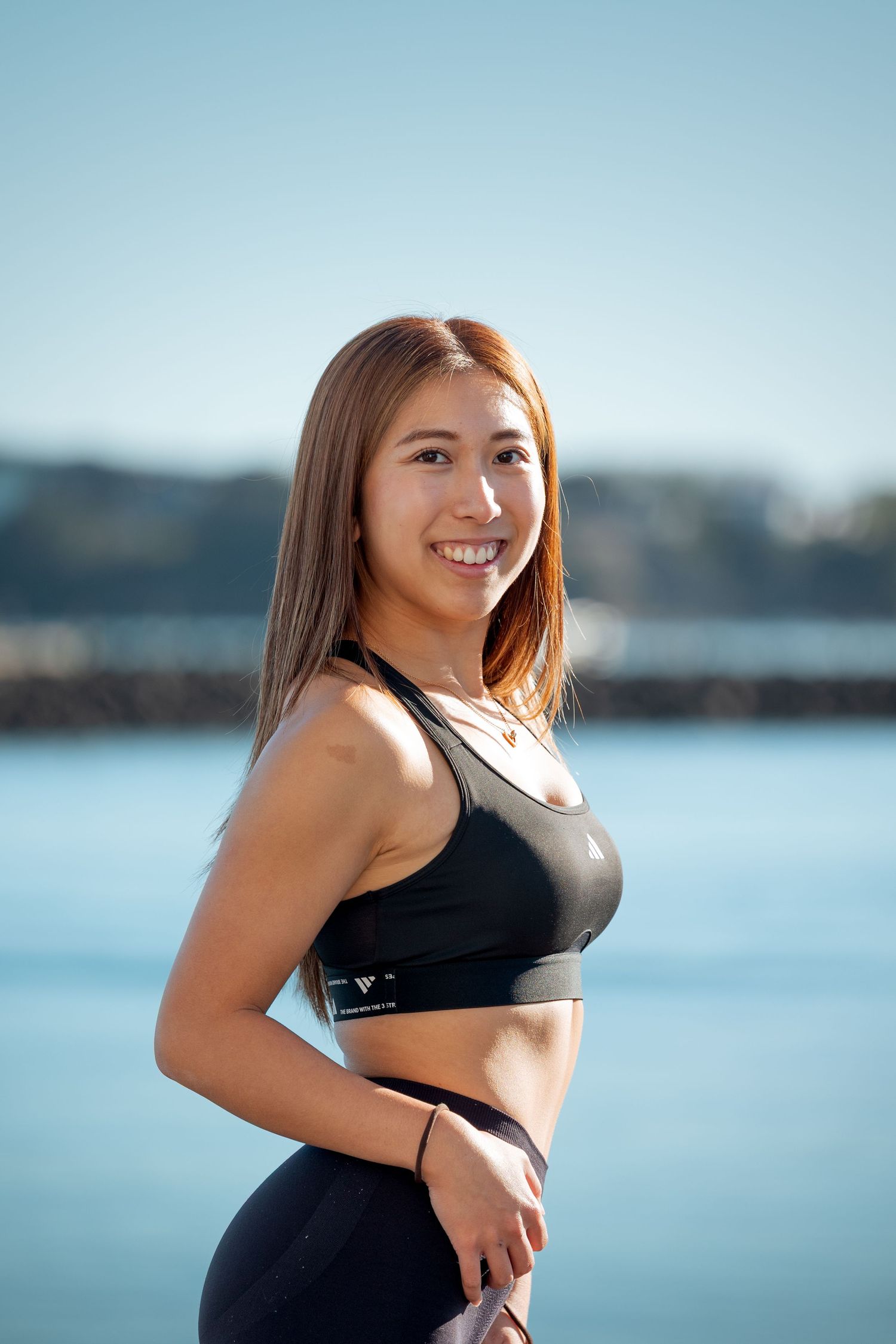 Smiling Asian woman in black Adidas sports bra and leggings posing by a waterfront on a sunny day.
