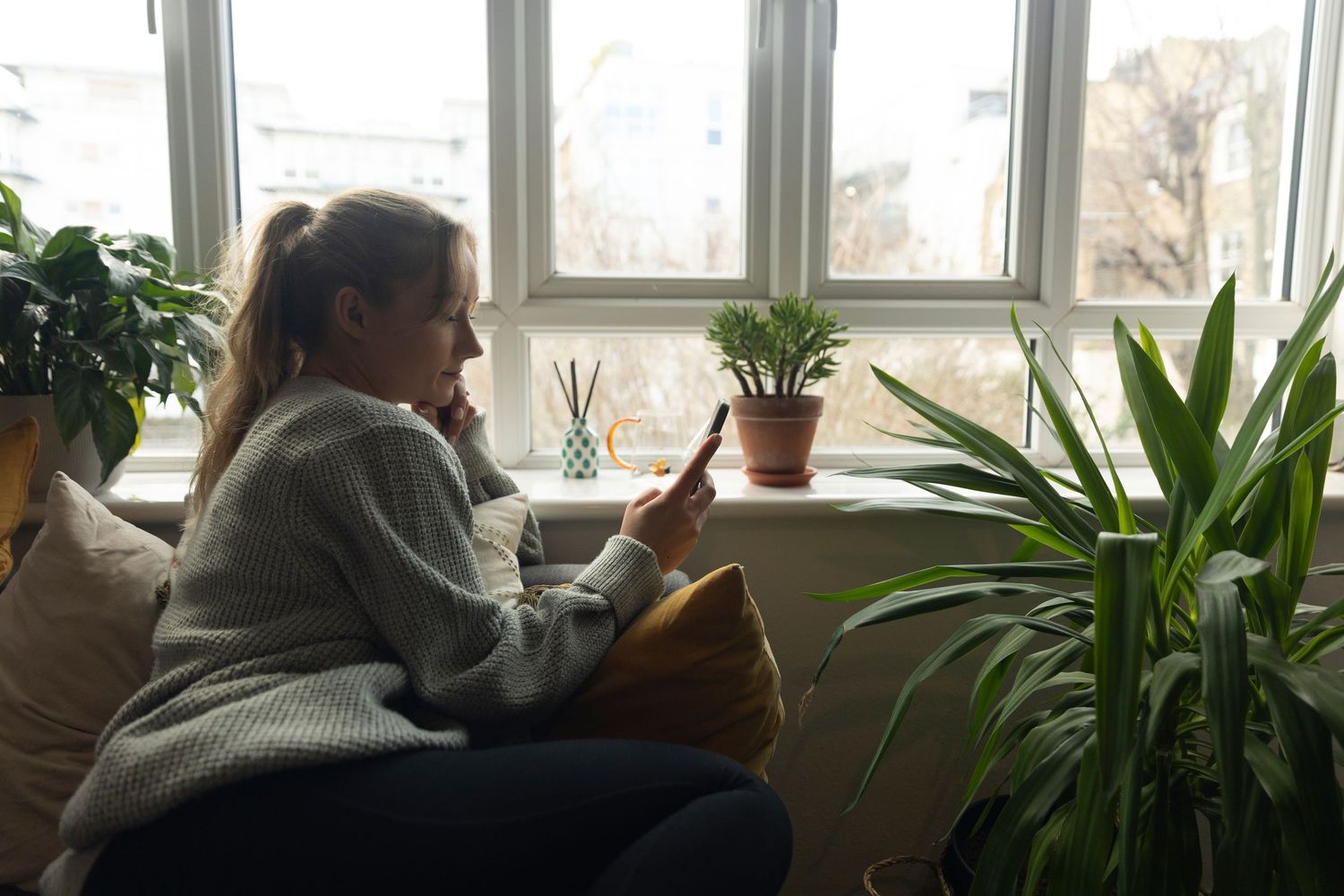Woman in gray sweater sitting on couch using smartphone near window surrounded by indoor plants.