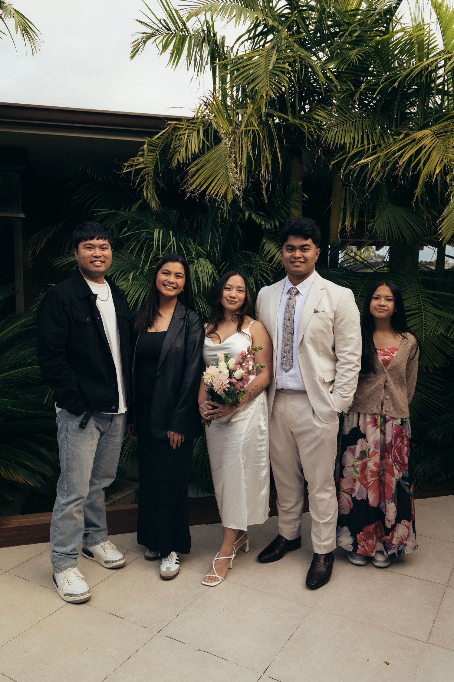 Group of five people posing outdoors at a wedding, with tropical palm trees in the background.