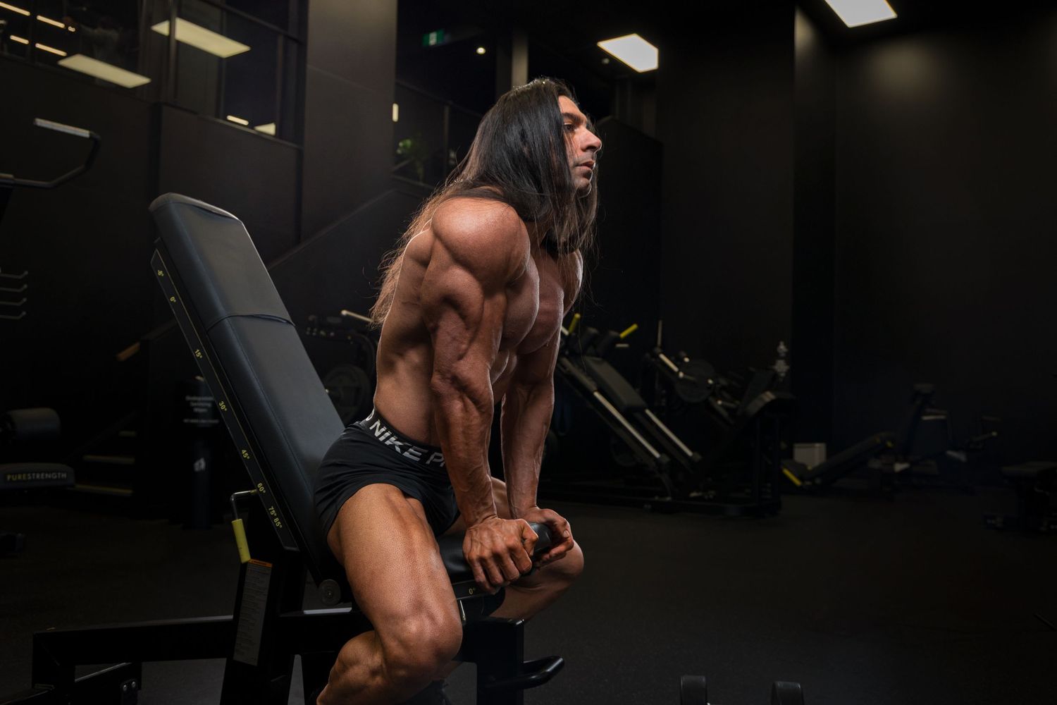 Muscular bodybuilder with long hair posing on a gym bench, showcasing defined back and arm muscles in a dark fitness center.