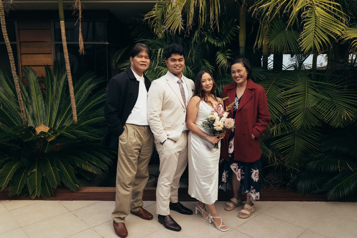 Four people posing together outdoors at a wedding, surrounded by tropical greenery, with the couple dressed in elegant attire.