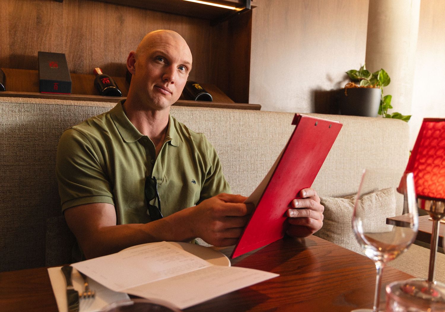 Bald man in green polo shirt reading a red menu at a restaurant table with wine glasses.