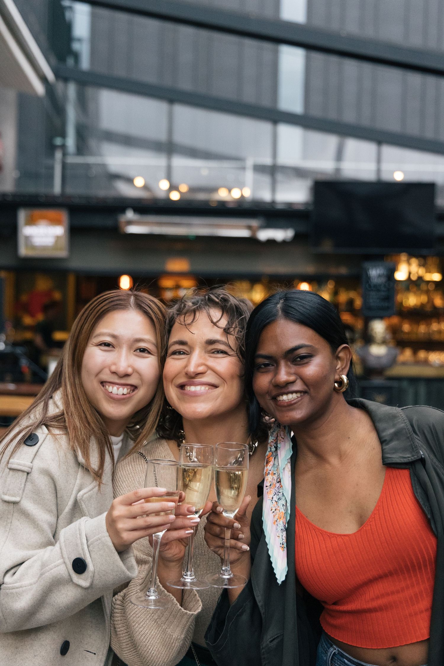Three diverse smiling women holding champagne glasses together outside a restaurant.