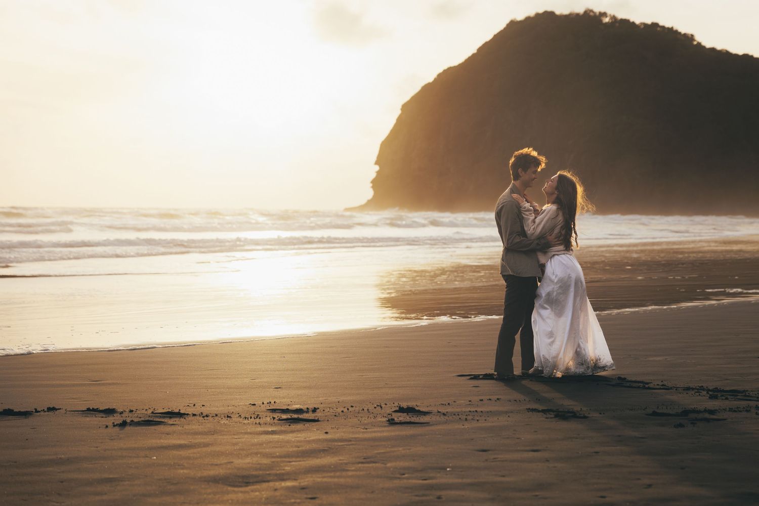 Couple embracing on a sandy beach at golden sunset with ocean waves and rocky headland in background.