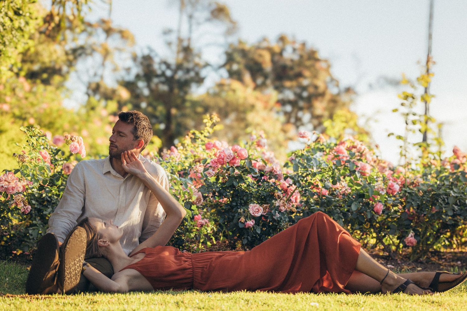 Romantic couple relaxing on green grass beside vibrant pink rose bushes in a sunny garden setting.